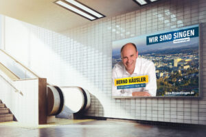 Großflächenplakat „Wir sind Singen“ mit Bernd Häusler in einer Unterführung neben einer Rolltreppe