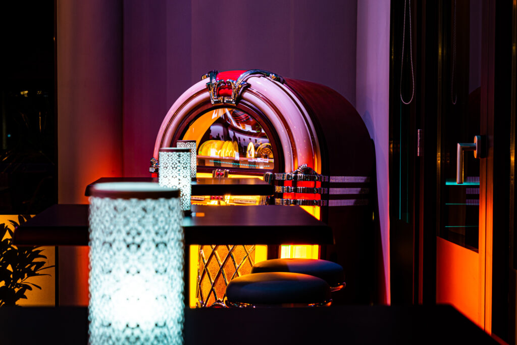 Beleuchtete Retro-Jukebox in einer Bar, davor Tisch mit Tischleuchten und Barhockern